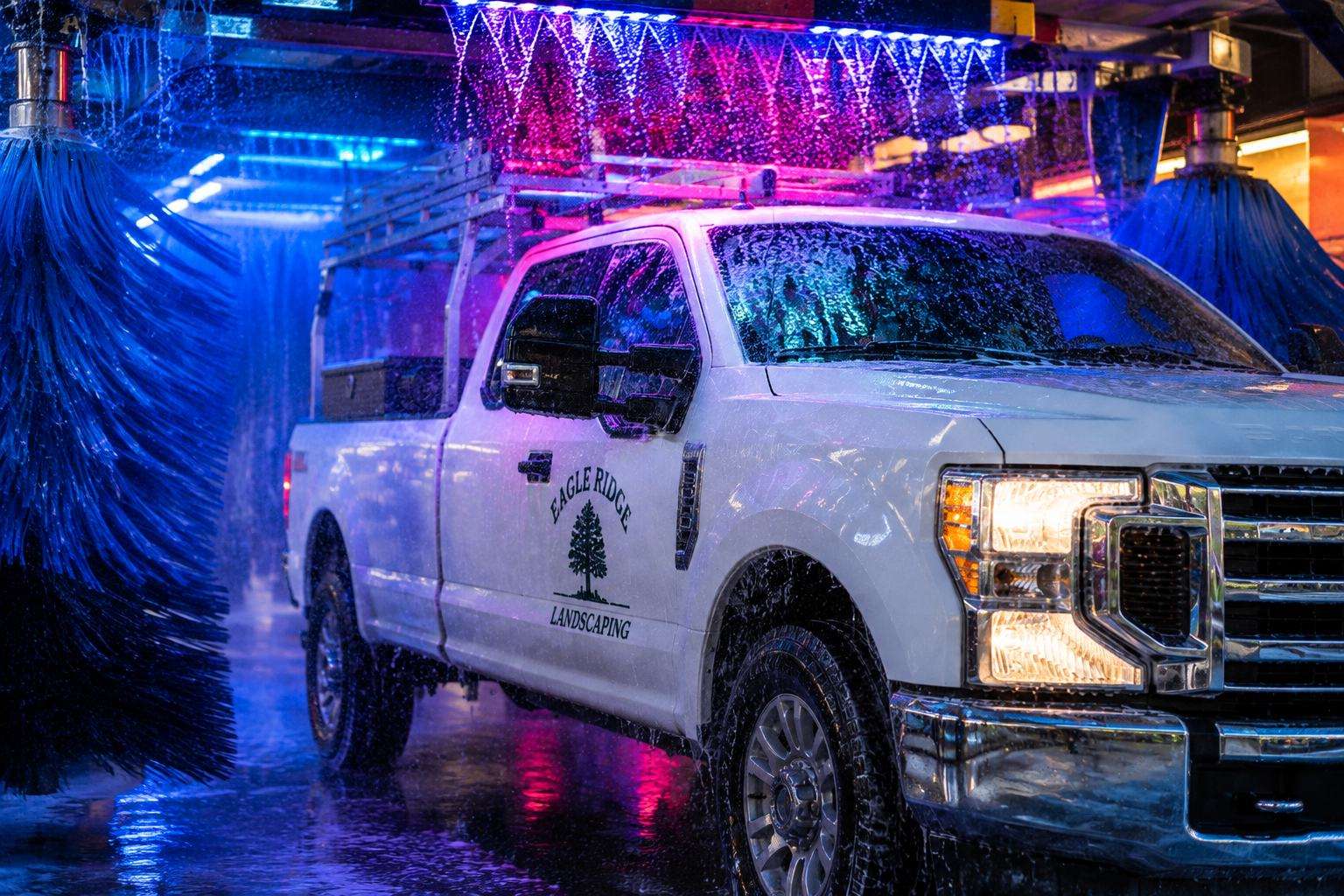 White work truck with a ladder rack rolling through the wash tunnel under blue and pink lights