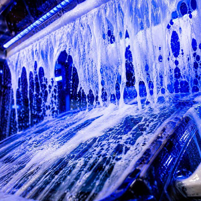 Curtain of white foam pouring over a car roof under blue lights inside the wash tunnel